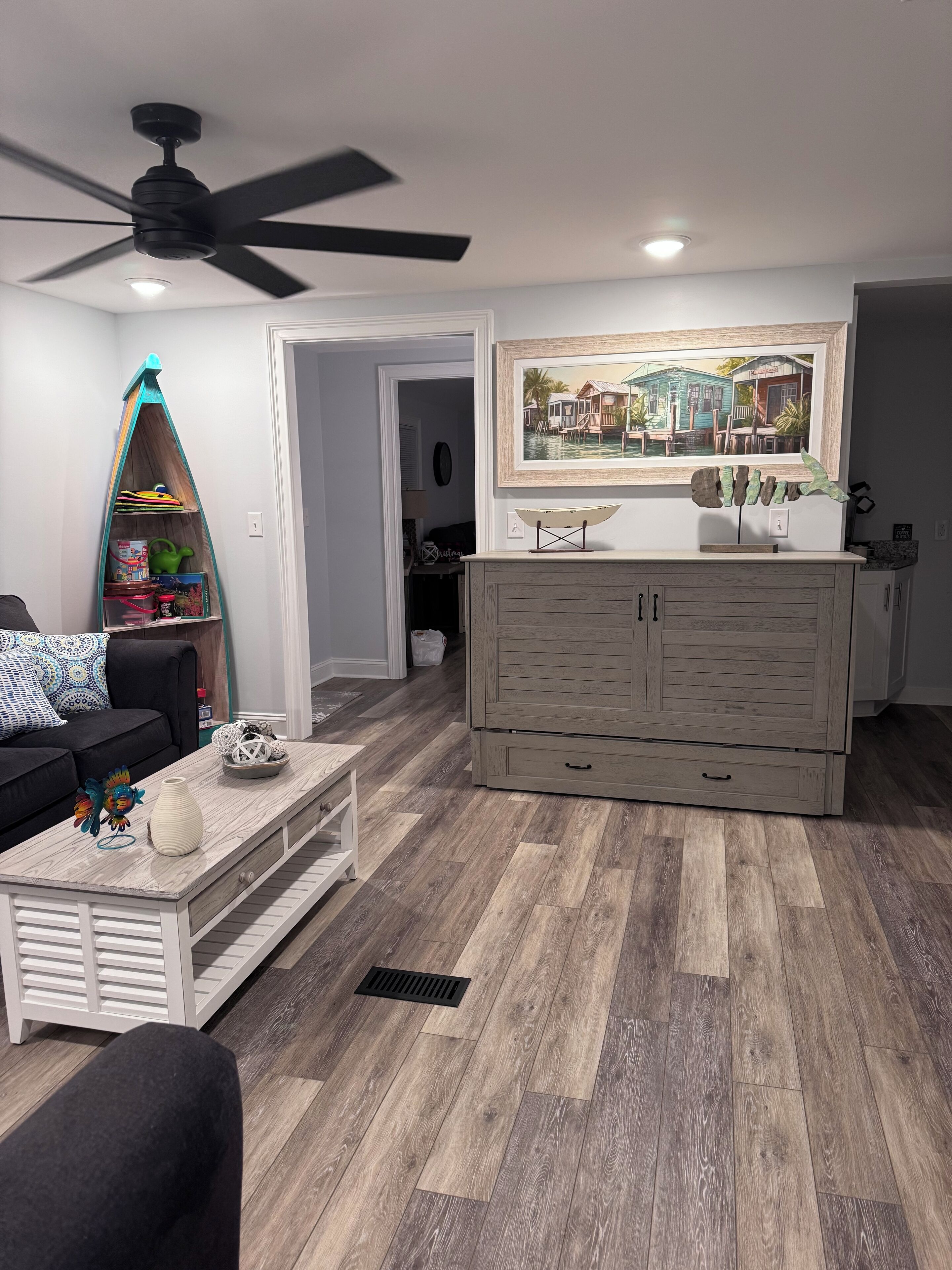 Un salon doté d’un ventilateur de plafond, un coin repas avec un buffet en bois, et un tableau représentant un paysage marin accroché au mur.