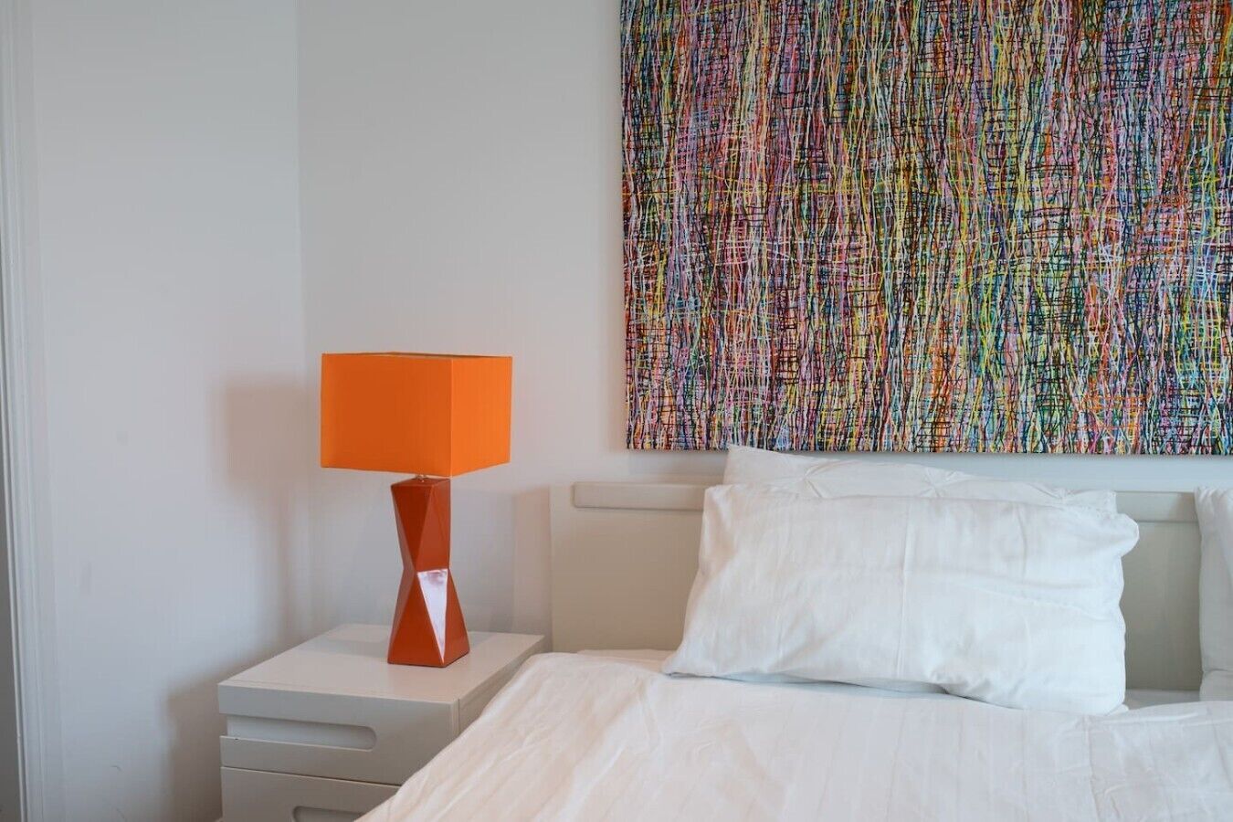 Une chambre d’hôtel avec un lit, du linge de lit blanc, une table de chevet avec une lampe et une peinture abstraite aux couleurs vives accrochée au mur.