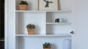 A white shelving unit with potted plants, a framed picture, and a white device on the lower shelf.