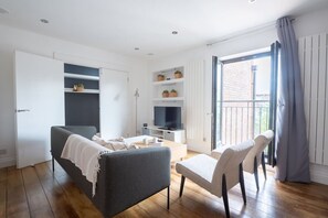 A modern living room with a grey sofa, a small table, a TV, and a balcony with a view. - King Bed, Full Kitchen | Blackout Curtains (London)