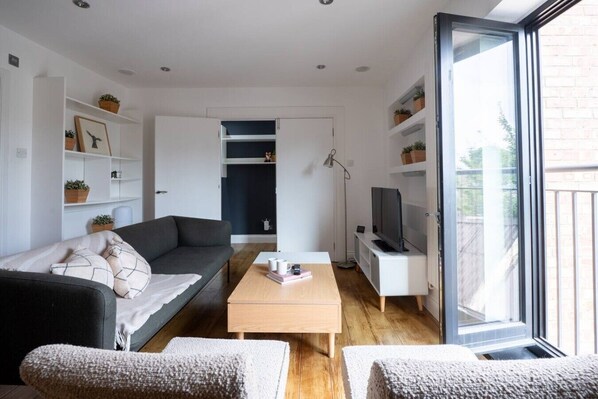 Una sala de estar moderna con sofá, mesa de centro y televisor, un puerta corrediza de vidrio hacia el exterior y estanterías empotradas con plantas.