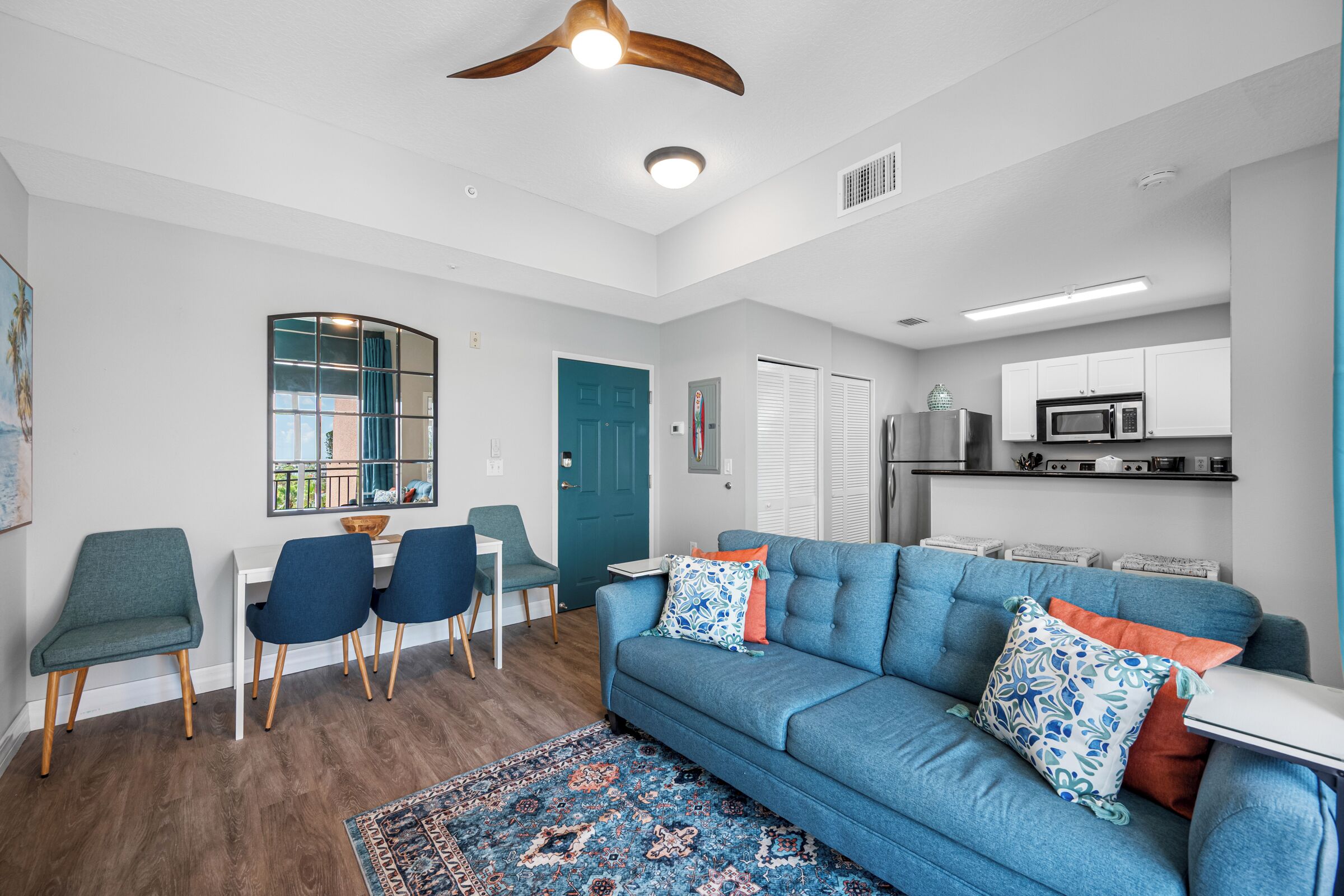 A modern living room with a blue sofa, a dining area with chairs, a kitchen with a microwave, and a ceiling fan.