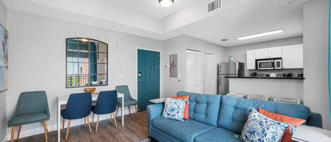 A modern living room with a blue sofa, a dining area with chairs, a kitchen with a microwave, and a ceiling fan.