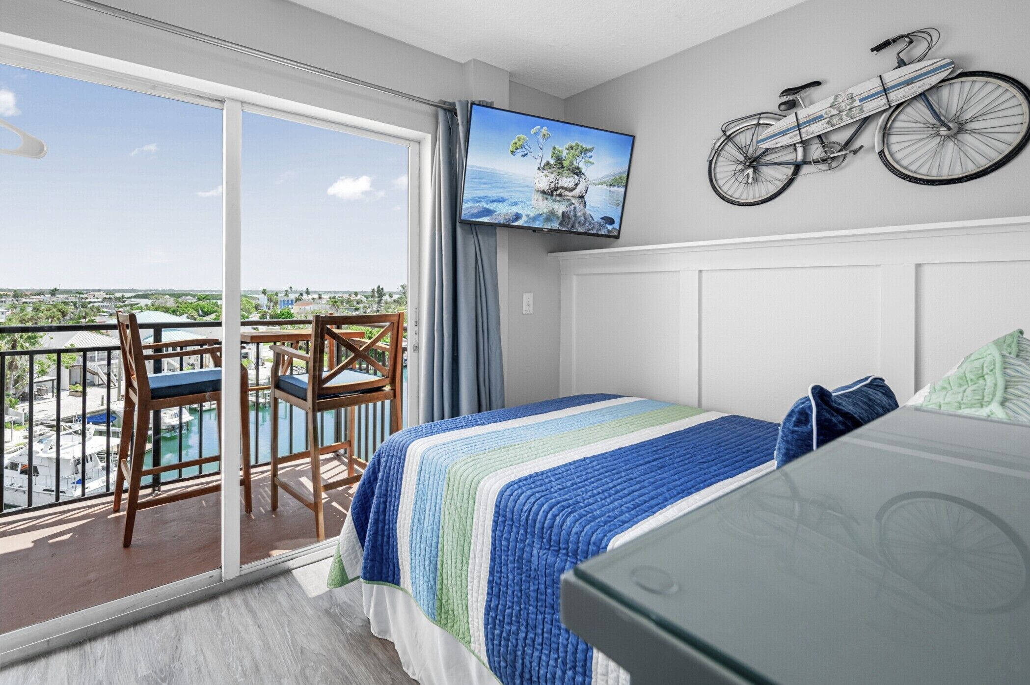 Ein Schlafzimmer mit Balkon, einem Bett mit gestreifter Decke, einem Wand-Fernseher und einer Fahrrad-Dekoration an der Wand.