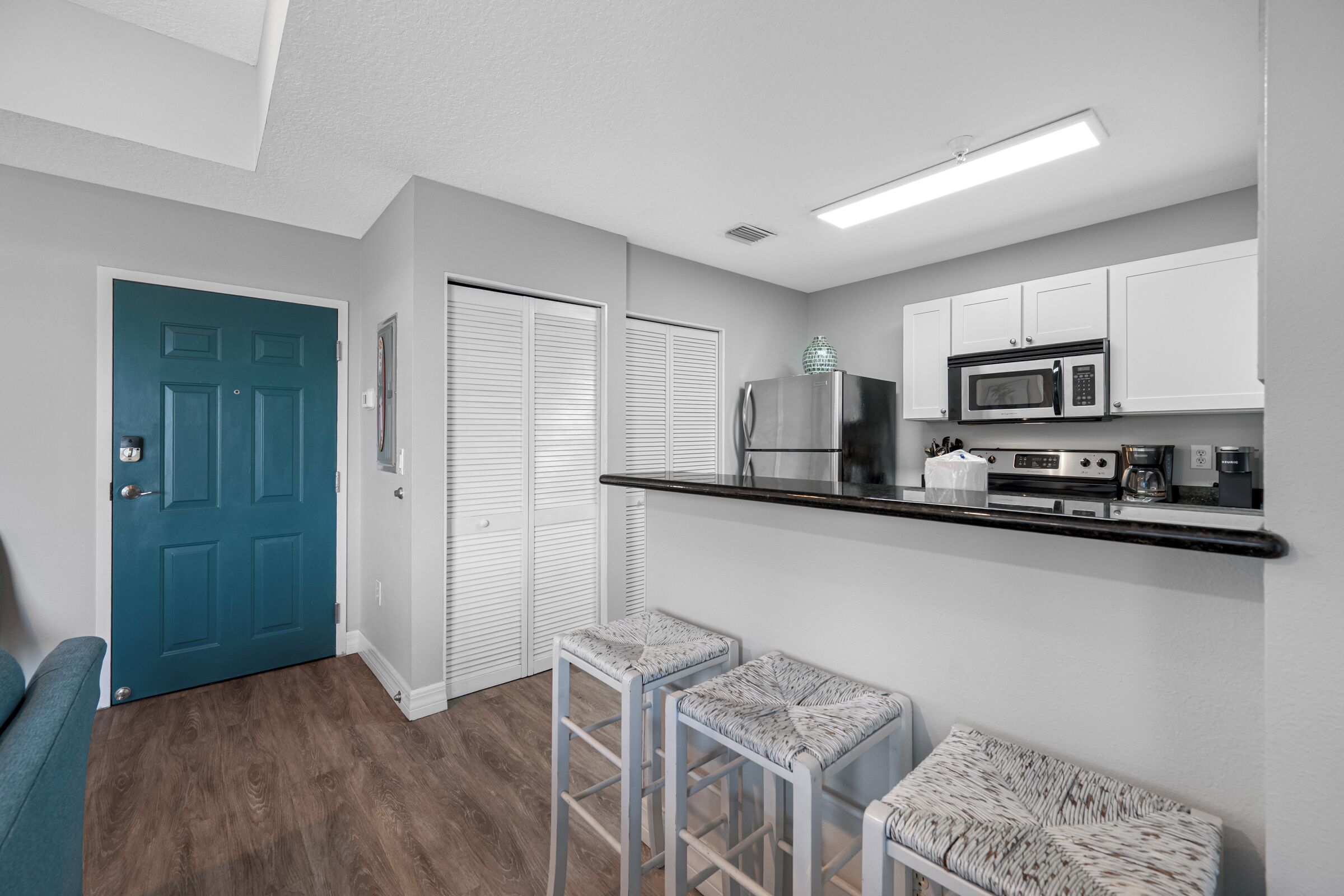 A compact kitchen with white cabinets, a microwave, and a coffee maker. There are two stools and a bench.