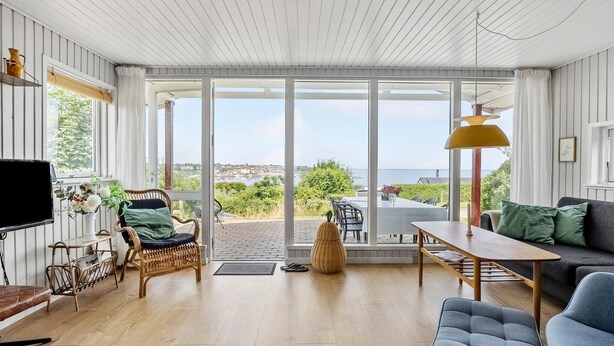 Un vaste salon doté d’une grande porte-fenêtre coulissante donnant sur une terrasse, d’un plancher en bois et offrant une vue sur la mer.