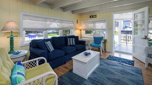 Una sala de estar acogedora con un sofá azul oscuro, una mesa de centro blanca y un sillón de mimbre. Hay dos lámparas sobre mesas auxiliares, y una puerta que da al exterior.