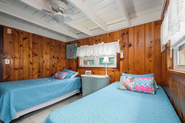 A room with two beds, wooden walls, a ceiling fan, a window with lace curtains, and a small table with a lamp.