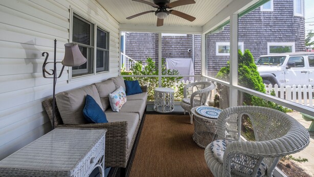 A covered porch with a ceiling fan, wicker furniture, and a view of a white picket fence and parked cars.