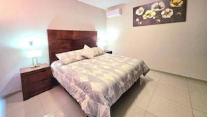 A bedroom with a bed, nightstands, a wall-mounted air conditioner, and a floral painting.