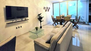 A modern living room with a flat-screen TV, a sofa, a glass coffee table, and a dining area with chairs and a table.