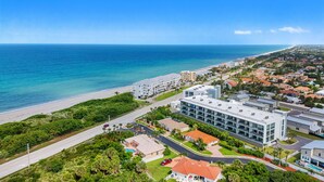 Vue aérienne d’une zone côtière comprenant une plage, des immeubles résidentiels et un hôtel moderne doté d’un toit plat et de plusieurs étages.