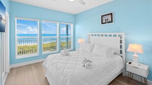 Une chambre à coucher comprenant un lit, deux tables de chevet, un ventilateur de plafond et une fenêtre avec des stores.