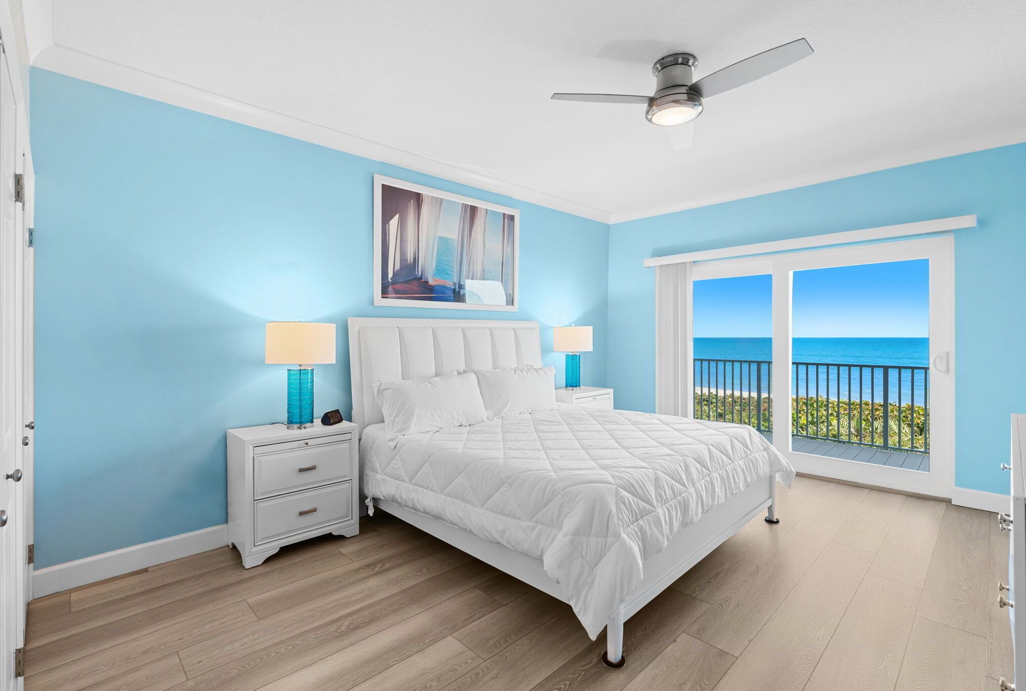 A bedroom with a white bed, two bedside tables with lamps, a ceiling fan, and a large window with a view of the ocean.