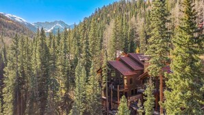 A log cabin with multiple levels, surrounded by dense forest and mountains in the background. - Apex - Walk to Trails + Hot Tub | Sauna & Theater (Mountain Village)