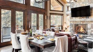 A spacious living area with a fireplace, large windows, and a dining table set for a meal. - Apex - Walk to Trails + Hot Tub | Sauna & Theater (Mountain Village)