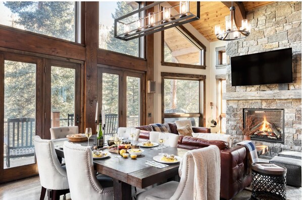 A spacious living area with a fireplace, large windows, and a dining table set for a meal. - Apex - Walk to Trails + Hot Tub | Sauna & Theater (Mountain Village)
