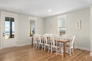 A dining area with a wooden table and white chairs, large windows with blinds, and a view of the beach.