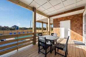 Un espacio al aire libre techado con una terraza de madera, una mesa con sillas y vista a una piscina y una casa al fondo.