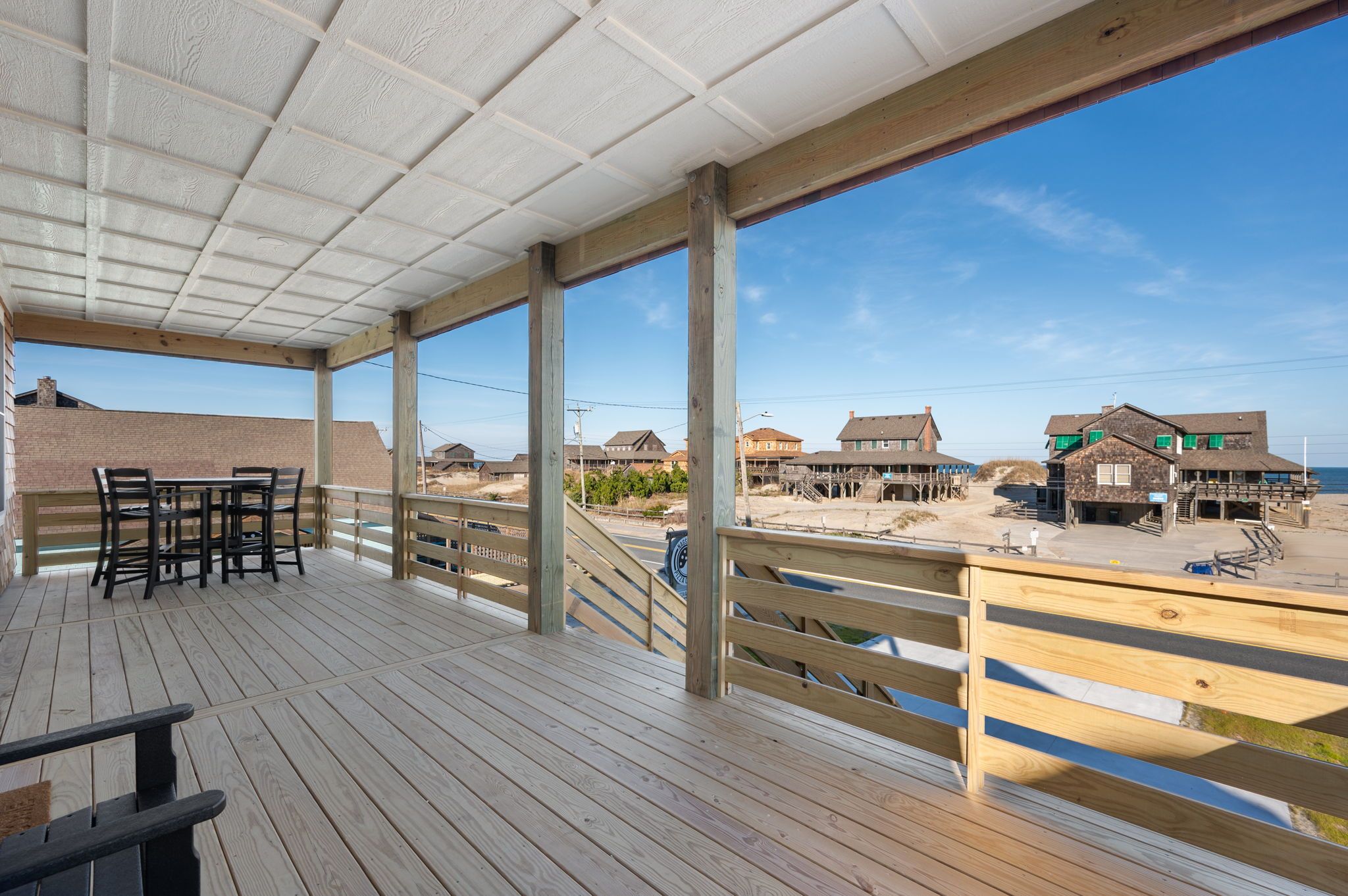 A wooden deck with a table and chairs, overlooking a beach with houses and a clear sky.