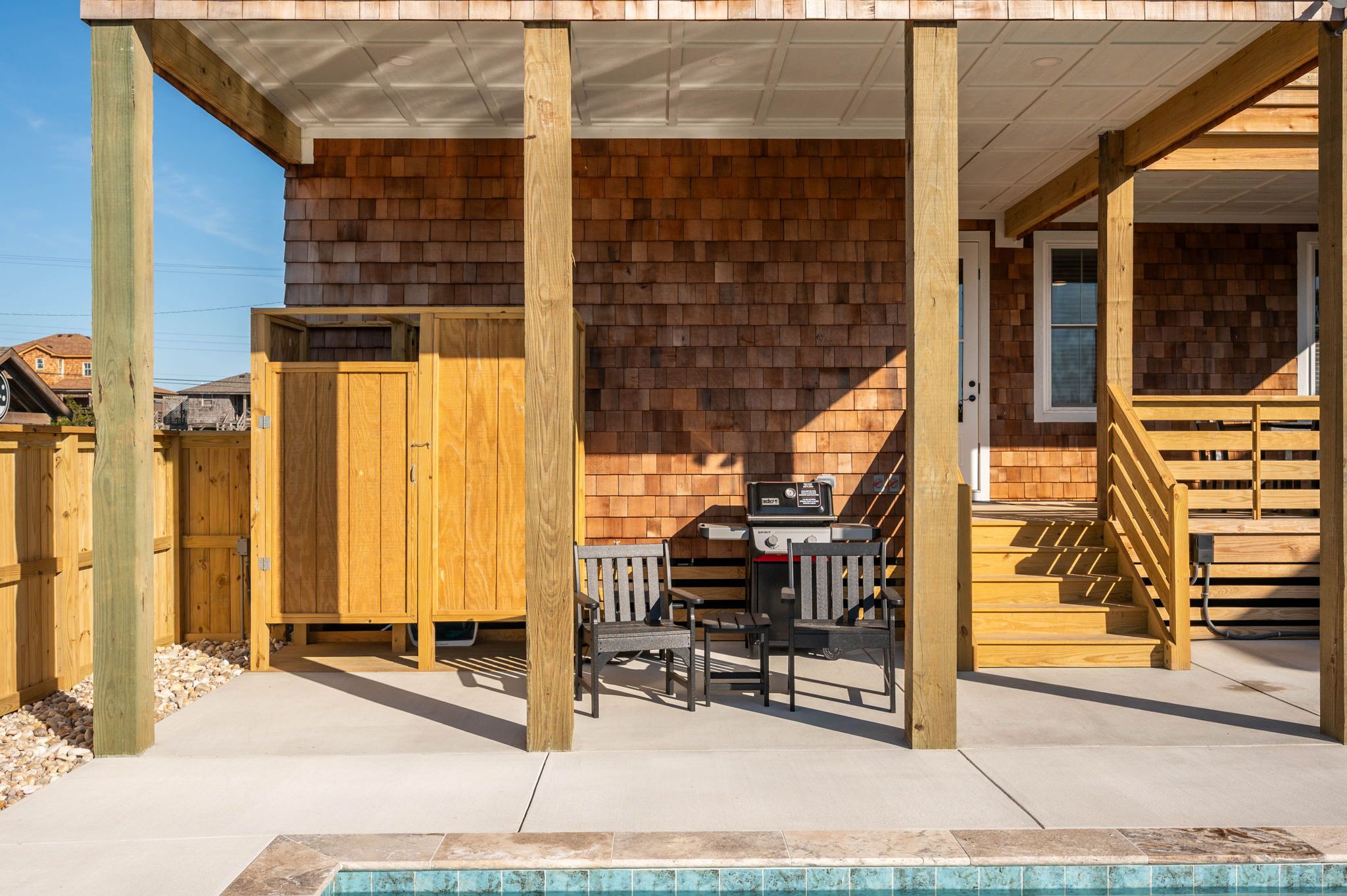 Eine überdachte Terrasse mit Grill, Holzstühlen und einer Holztreppe zu einer Tür.