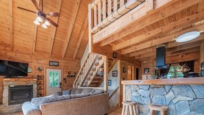 A wooden cabin interior with a stone fireplace, a sofa, a bar area, and a staircase.