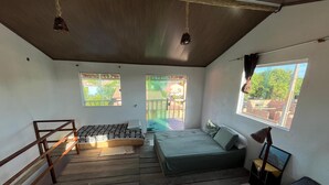 A compact room with a wooden floor, two beds, a small table, and a window with a view of a building and trees. - Hospedaria Labelle - Xandó Caraíva (Porto Seguro)