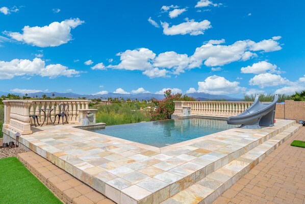 Una piscina con tobogán, baldosas de piedra y vistas a montañas.