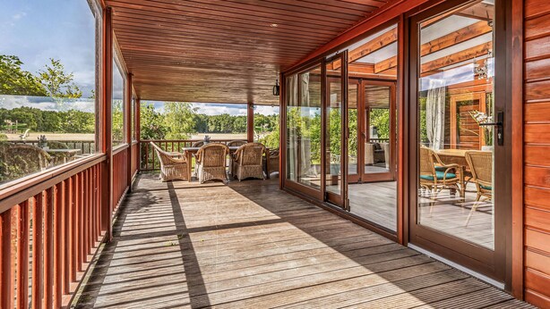 Una terraza de madera con zona cubierta, mobiliario de mimbre y grandes puertas correderas que dan a un espacio al aire libre.