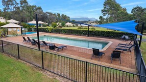 Una piscina rectangular con sillas de descanso y sombrillas, rodeada por un patio de ladrillo y una cerca de metal negro.