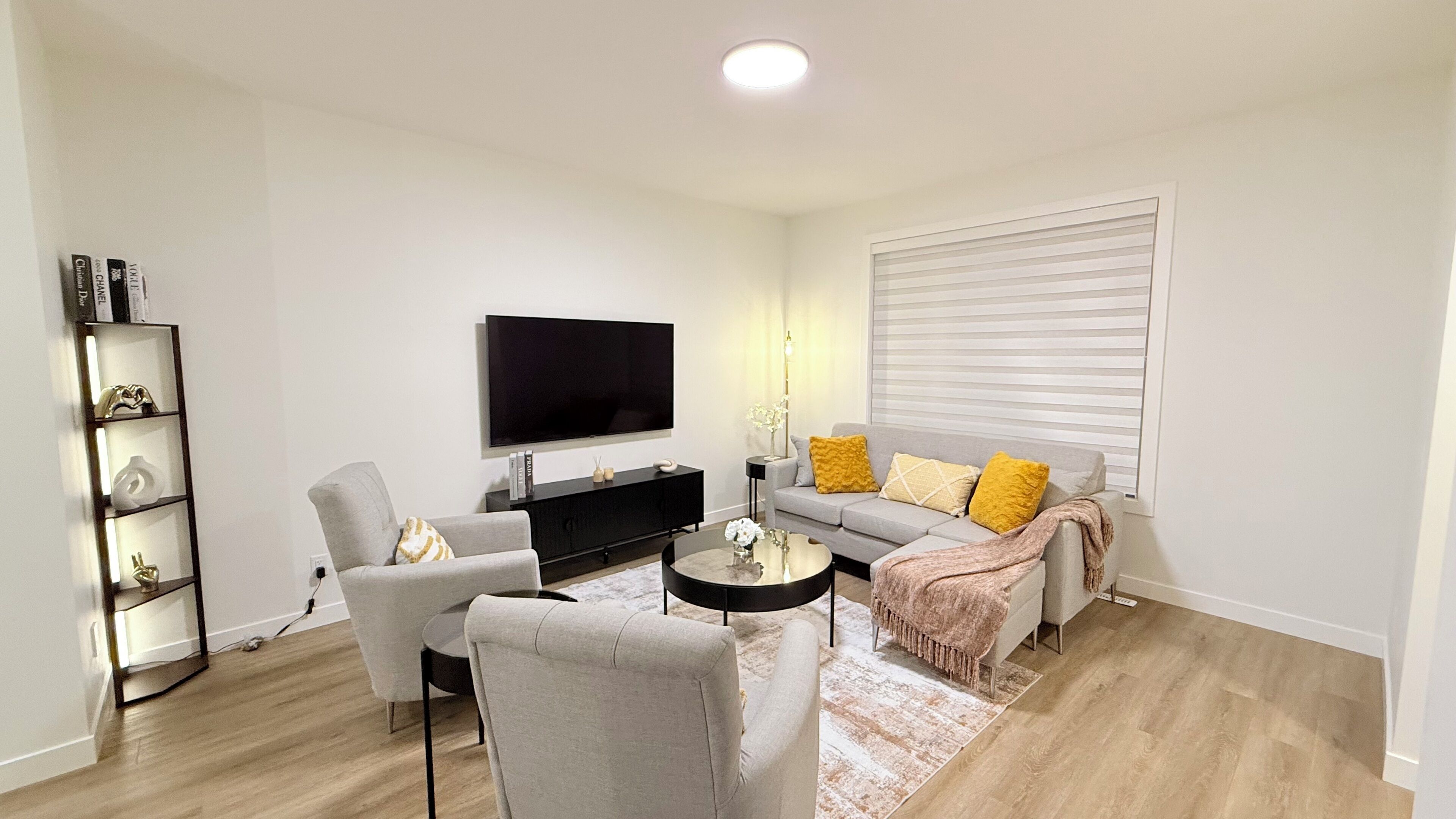 Een moderne woonkamer met een flatscreen televisie, een grijze bank, fauteuils, een salontafel en een boekenplank.