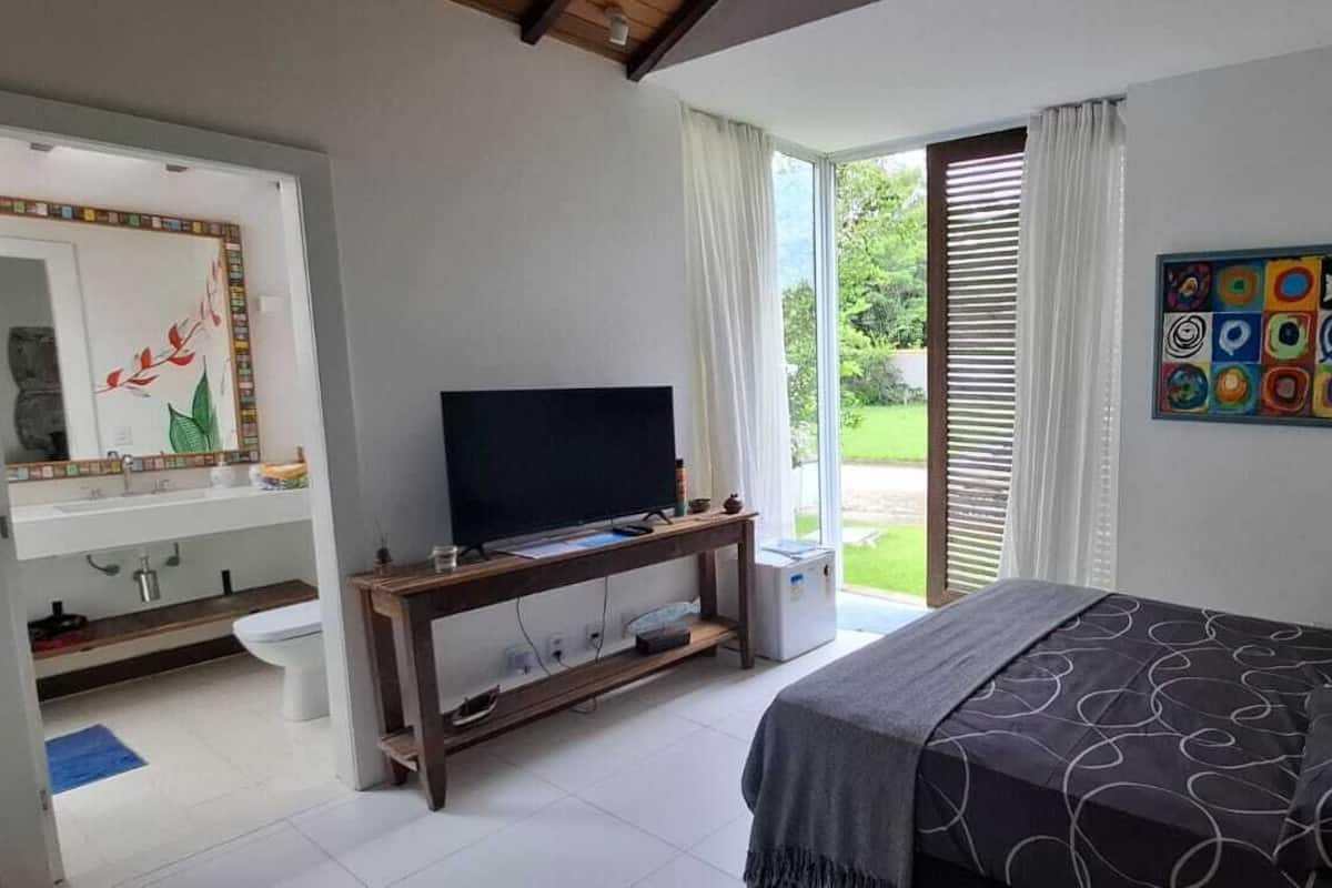 A bedroom with a bed, a television on a wooden stand, a bathroom with a mirror, and a colorful abstract painting on the wall.