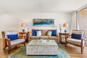 A living room with a beige sofa, two wicker chairs, a coffee table, and a painting of a blue seascape.