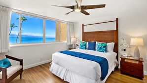 A bedroom with a large bed, a ceiling fan, a nightstand, a lamp, and a view of the ocean.