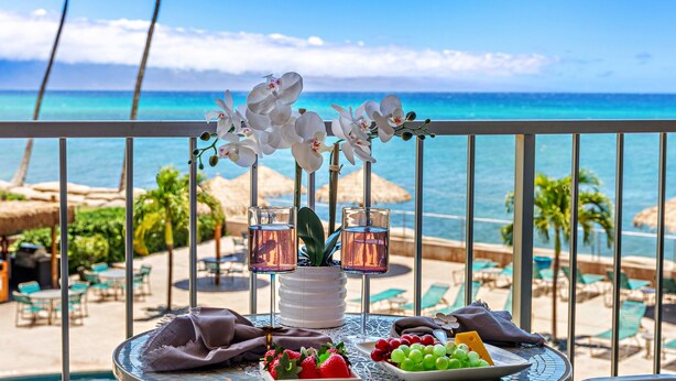 A table with a white vase of orchids, two glasses of pink drink, and a plate of fruit on a balcony overlooking a beach.