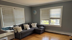 A living room with a sectional sofa, two windows with blinds, a small table, and a wicker basket. - Peaceful 2-bedroom apartment with AC in Spring Lake Heights close to the beach. (Spring Lake)