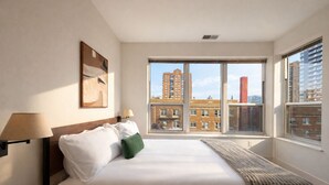A hotel room with a large bed, two bedside lamps, a view of buildings through a large window, and a painting on the wall. - Upscale Apartment Near Lake Michigan · Sleeps 6-7 (Milwaukee)