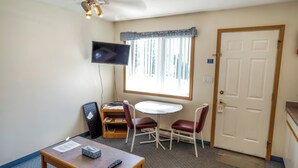 A small room with a TV mounted on the wall, a round table with two chairs, a desk with a chair, and a door with a sign. - Blue River Motel (Blue River)