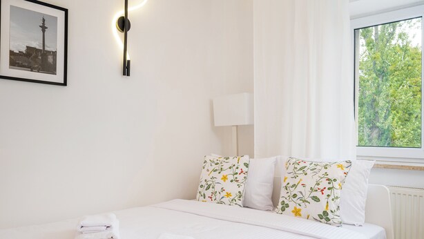 Un lit bien fait, avec du linge de lit blanc et des coussins à motifs floraux, un tableau encadré au mur et une fenêtre donnant sur un paysage verdoyant.