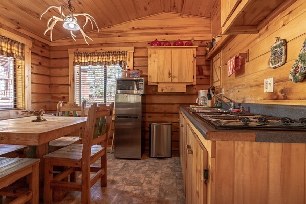 A wooden cabin kitchen with a dining table, chairs, a refrigerator, a stove, and a window with curtains.