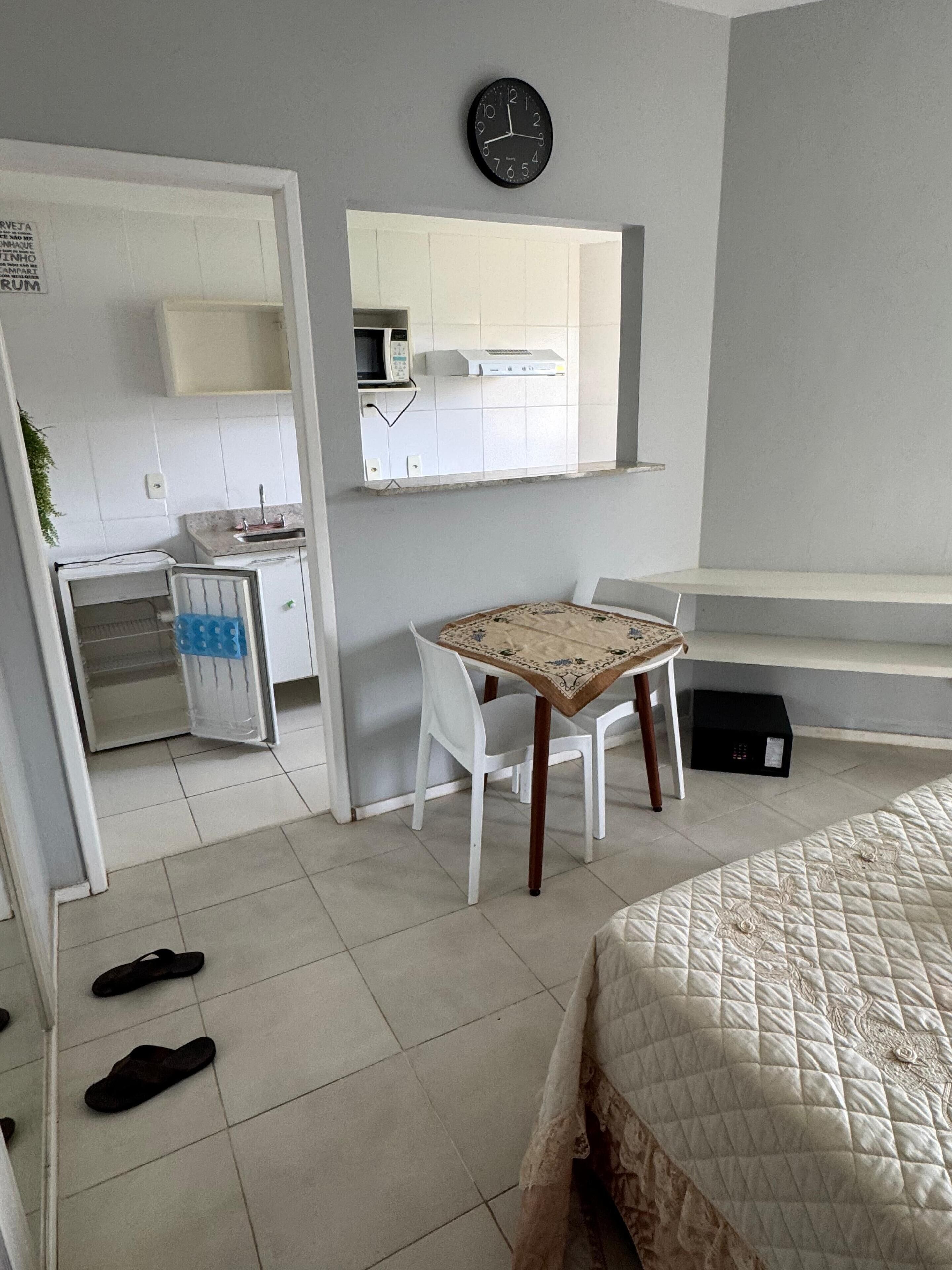 Een slaapkamer met een bed, een klein tafeltje, een stoel, een klok en een magnetron in de aangrenzende kitchenette.