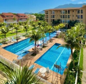 A swimming pool with multiple lanes, surrounded by palm trees and lounge chairs, with buildings in the background. - Wonderful loft in a condominium full of leisure activities and stunning beaches. (Sahy Mangaratiba)