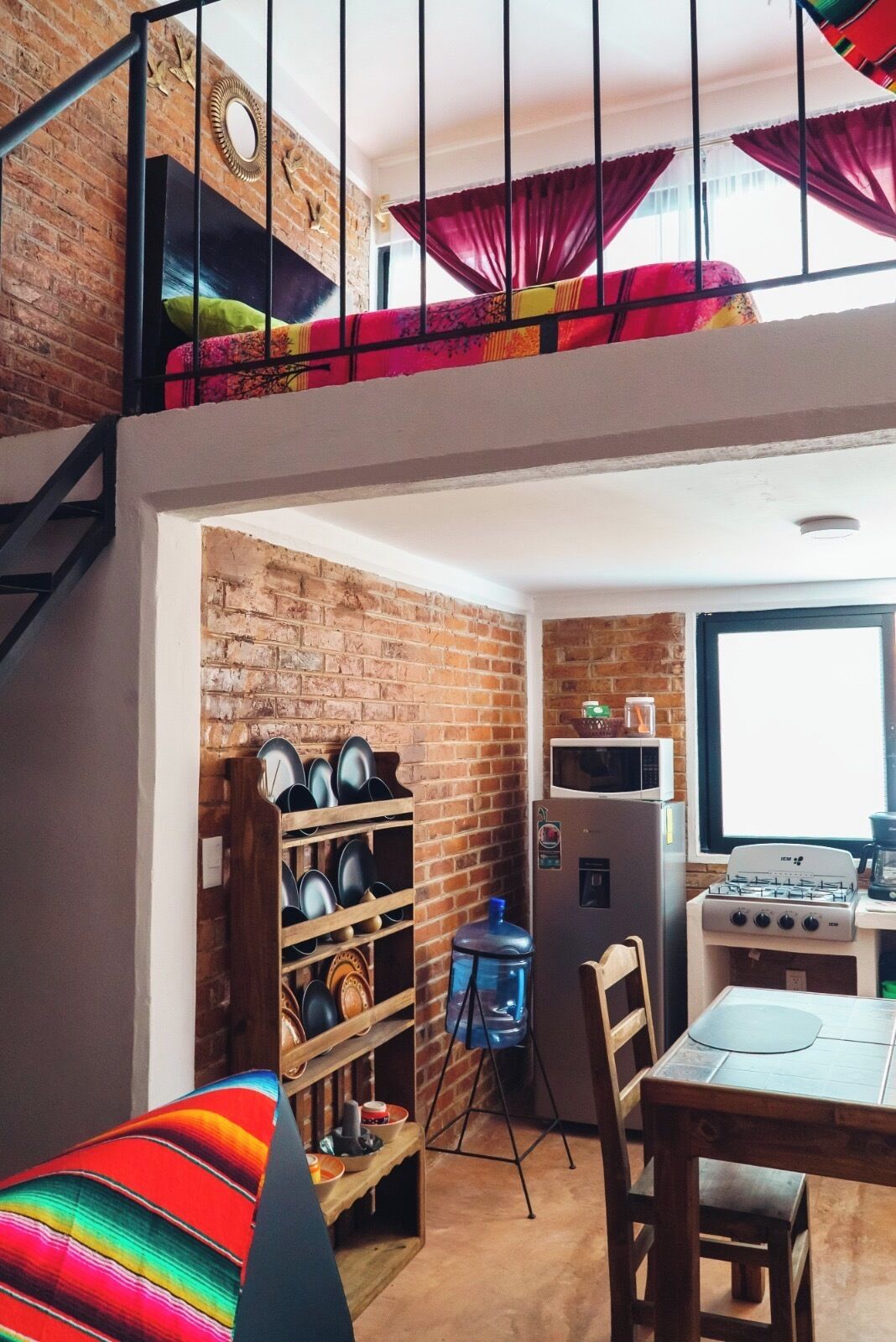 A loft-style room with a brick wall, a loft bed, a dining area with a table and chairs, a water cooler, and a kitchen with a stove and refrigerator.