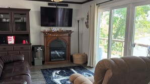 A living room with a fireplace, a ceiling fan, a TV, and a large window. - The Lakeview Cabin (Clarkson)
