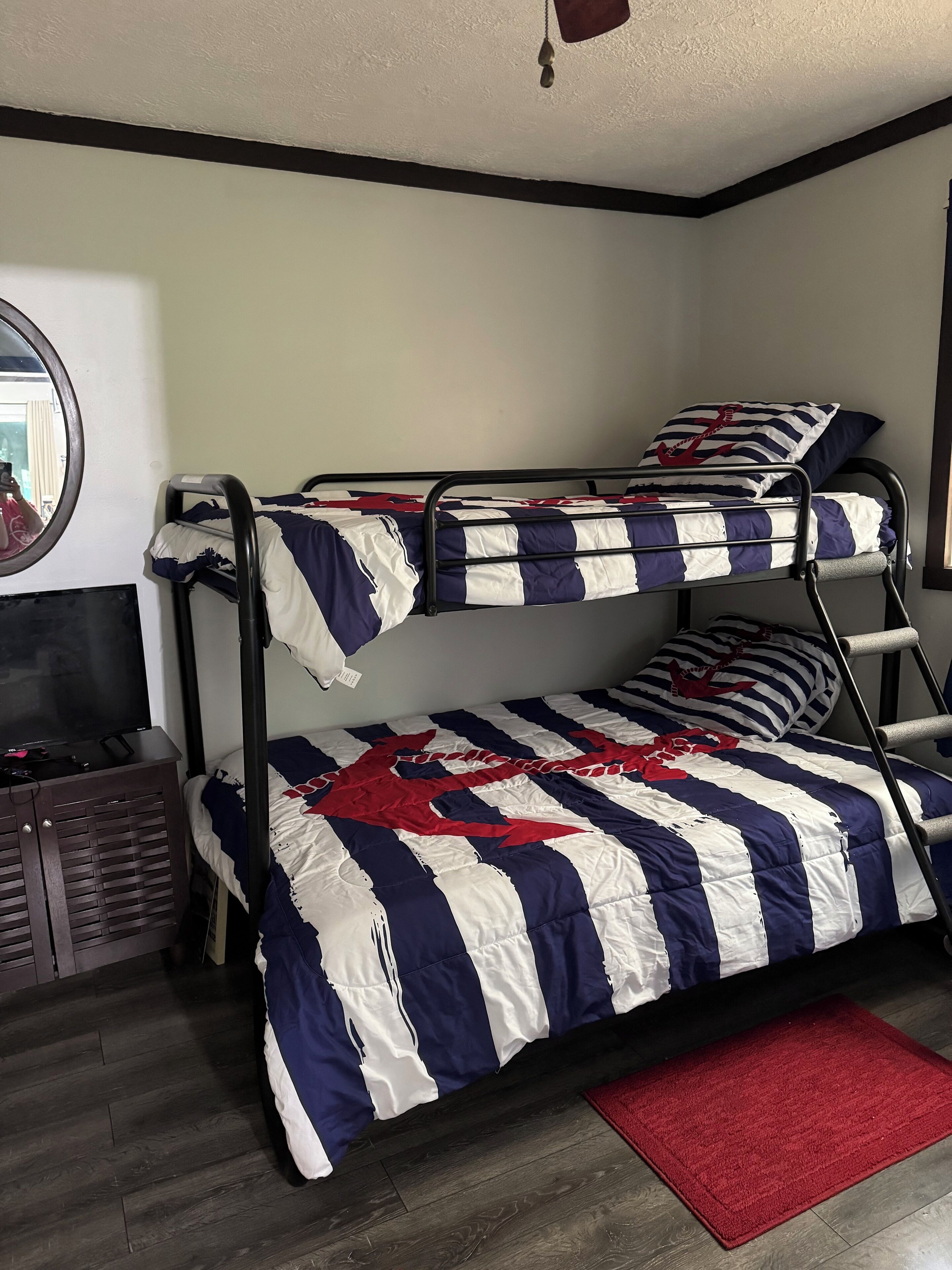 Un lit superposé avec une literie à rayures, un tapis rouge, un téléviseur posé sur un meuble en bois et un miroir au mur.