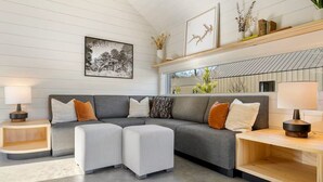A modern living room with a grey sectional sofa, two matching ottomans, a wooden coffee table, and a side table with a lamp. There is a framed black and white photograph on the wall and a shelf with decorative items above the sofa. - Charming 2-bedroom, lakeside cabin at Lake Cypress Springs in Mt. Vernon, TX (Mount Vernon)