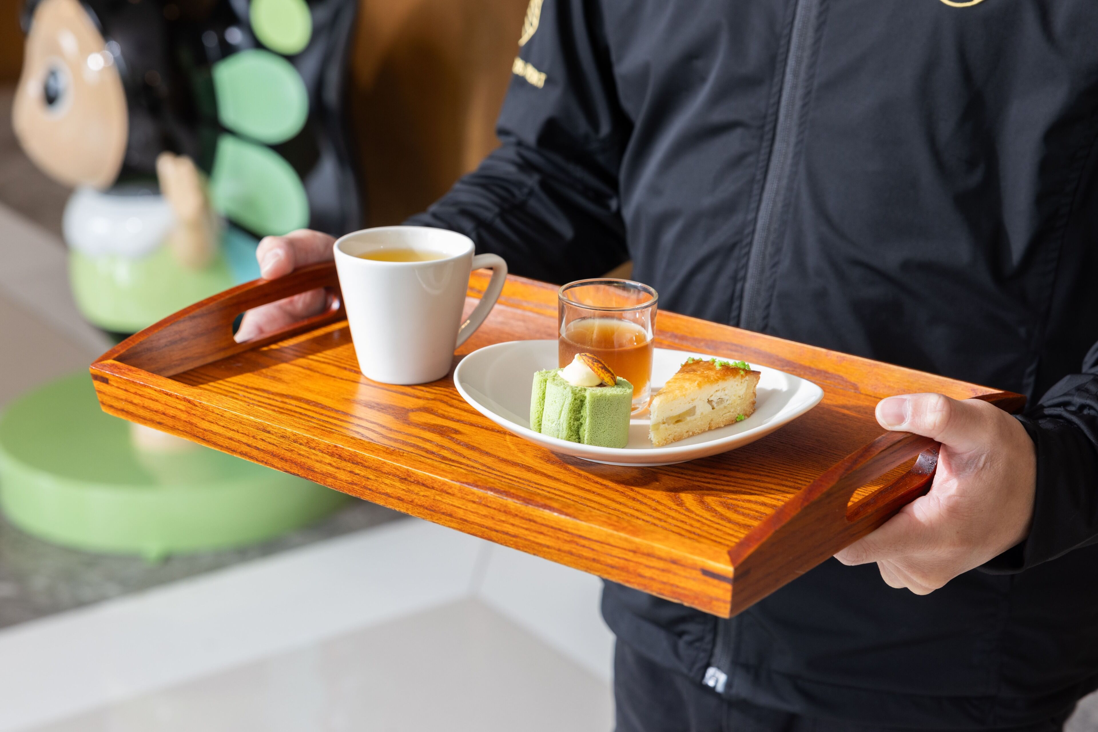 Una persona sostiene una bandeja con una taza de café, un vaso de té y un plato con un pastel.