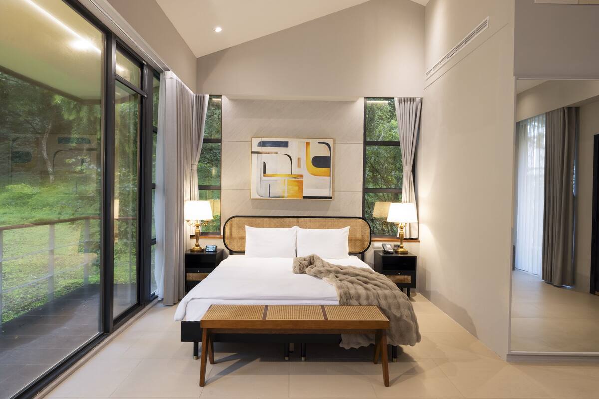 Habitación de hotel moderna con una cama grande, lámparas de noche, un banco y vistas a zonas verdes a través de amplios ventanales.