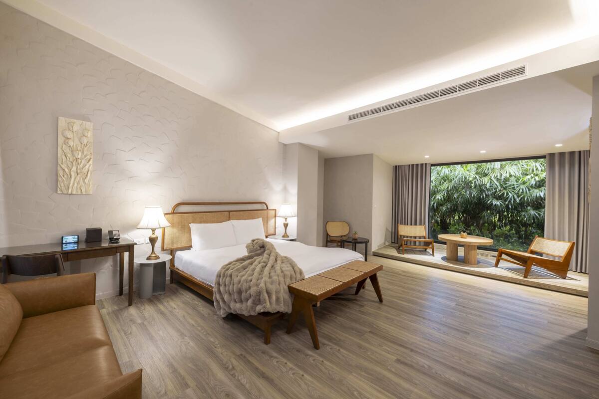 Habitación de hotel con cama, mesita de noche, lámpara, silla y vistas a zonas verdes a través de amplias ventanas.