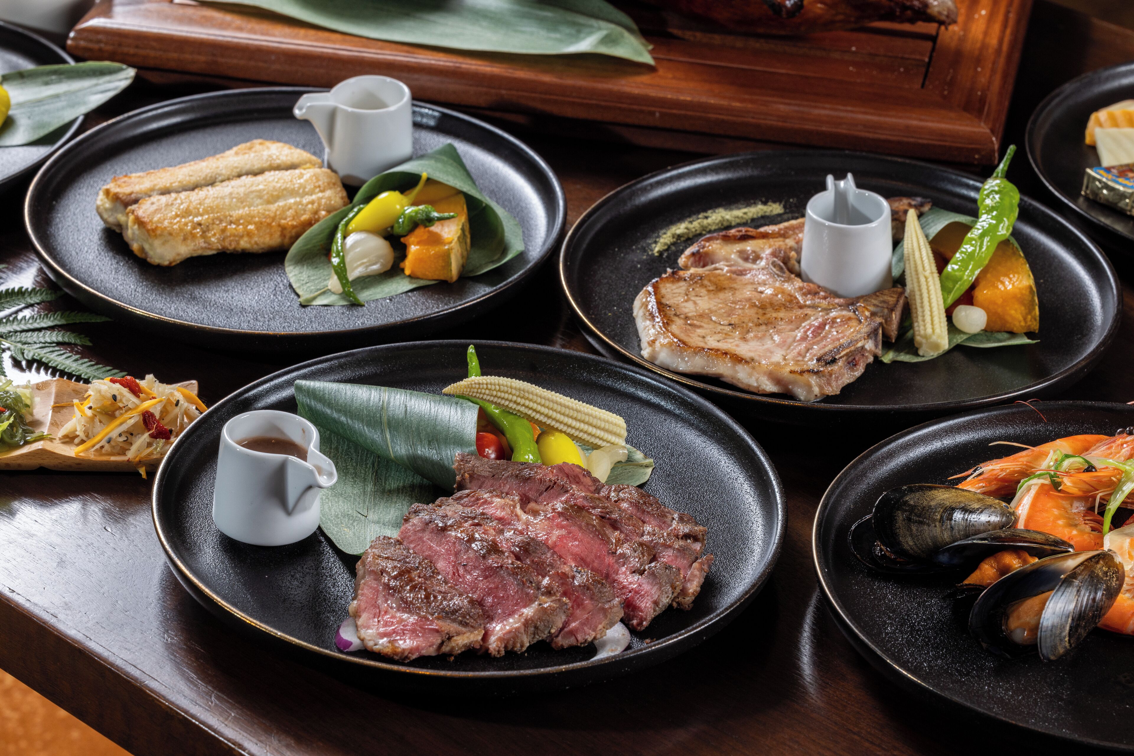 A variety of dishes on black plates, including steak, shrimp, and other seafood, with a small white cup on each plate.